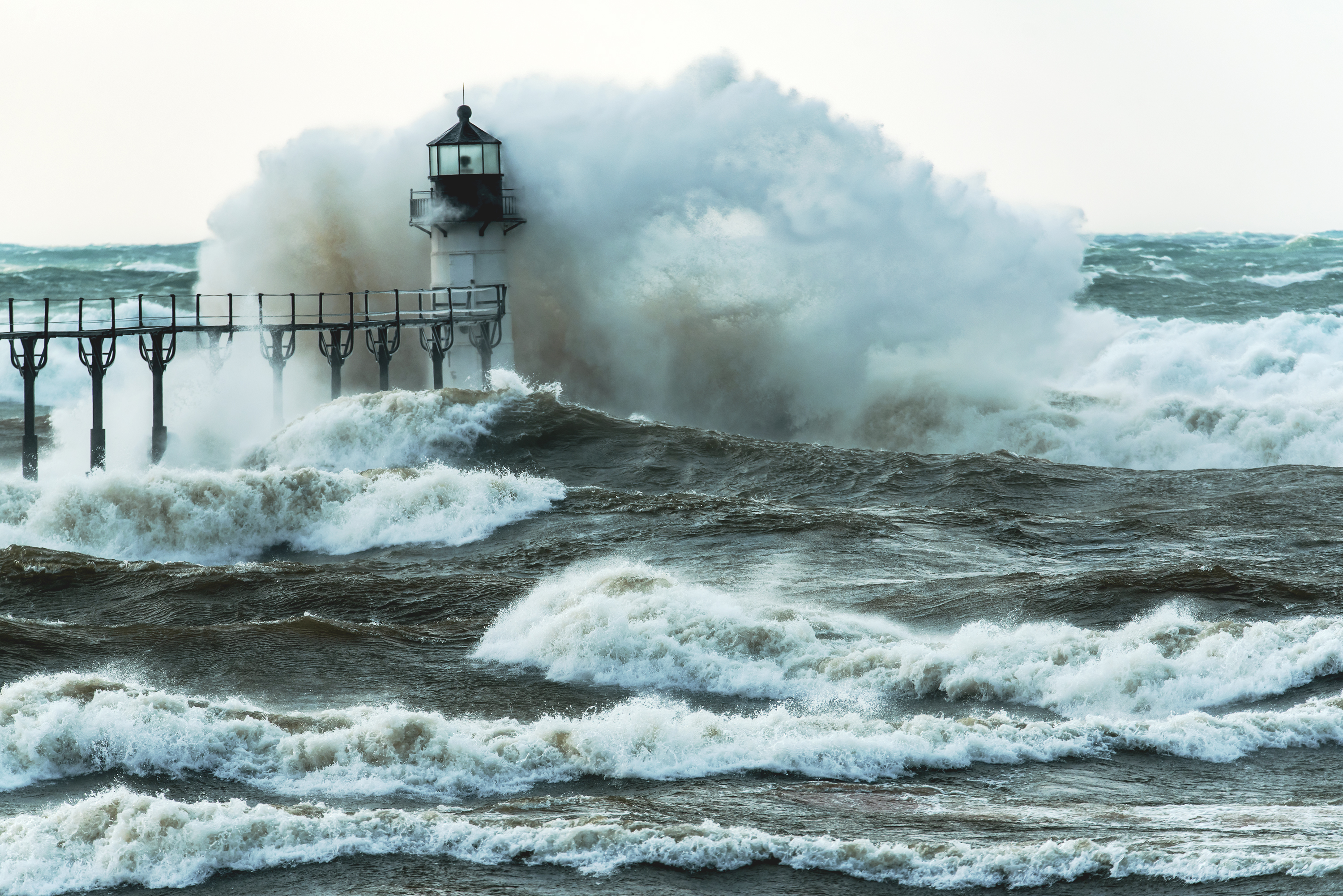 a large storm wave crashes over a lighthouse at the end of a pier