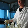 A scientist in a white lab coat stands in laboratory holding a jar of marine specimens. 