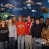 Students pose in front of an aquarium tank with fish.