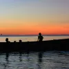 A silhouette of a small boat carrying 8 passengers on tranquil water, with a pink horizon. 