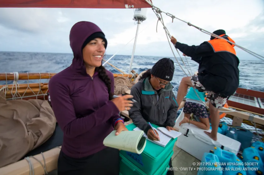 Four people working aboard the deck of a ship. Two are checking materials and noting observations while the two in the background adjust the canoe's rigging.
