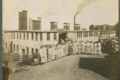 a group of women in period dresses from 1912 stand outside a factory with smoking stacks