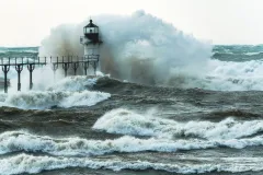 a large storm wave crashes over a lighthouse at the end of a pier
