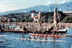 A sailing canoe sits in a Tahitian bay, surrounded by crowds of onlookers, and several smaller canoes full of people there to celebrate the occasion.