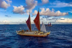 Two sailing canoes float on open water, with islands spotting the horizon. The canoe hulls are orange along the bottom half and white along the top, with brightly colored sails.