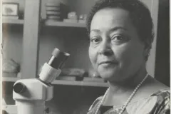 a woman with short black hair and a long necklace of pearls sits in front of a microscope and holds a small button coral