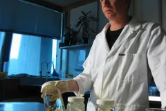 A scientist in a white lab coat stands in laboratory holding a jar of marine specimens. 