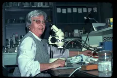 an older woman with short white hair smiles while sitting by a microscope