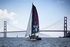 The Plastiki sailing vessel on the water with sails unfurled, in front of a large suspension bridge.