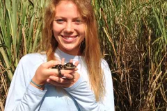 Alexis Temkin stands in marsh grass holding a reptile. 