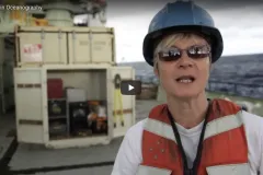 A screenshot from the video "Women in Oceanography" showing a woman standing on a research vessel. 