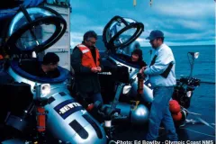two twin submersibles sit on a ship while the pilots ready for a mission