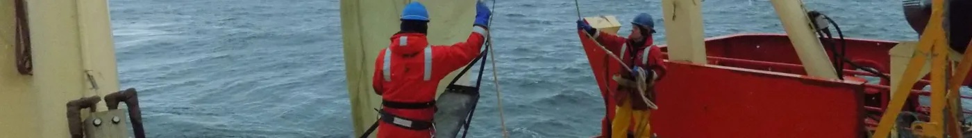 Two people in bright orange jackets work on a research vessel. 