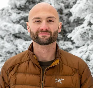 Andrew in profile wearing a brown puff jacket with snowy trees in the background