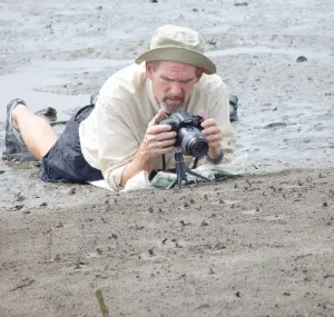 John Christy lies on the sand of a beach with a camera.
