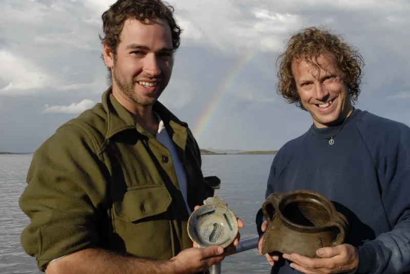 Two men each hold a ceramic vessel at the archaeological excavation