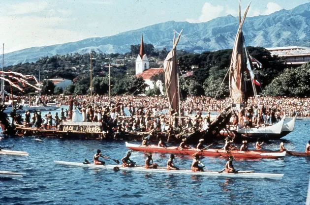A sailing canoe sits in a Tahitian bay, surrounded by crowds of onlookers, and several smaller canoes full of people there to celebrate the occasion.