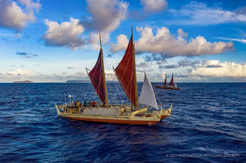 Two sailing canoes float on open water, with islands spotting the horizon. The canoe hulls are orange along the bottom half and white along the top, with brightly colored sails.