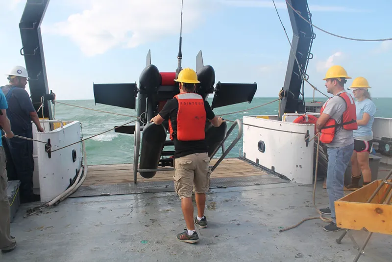 A large, black instrument is hauled onto a ship's deck by researchers wearing hard hats and life vests.