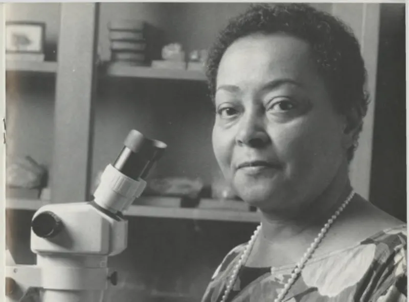 a woman with short black hair and a long necklace of pearls sits in front of a microscope and holds a small button coral