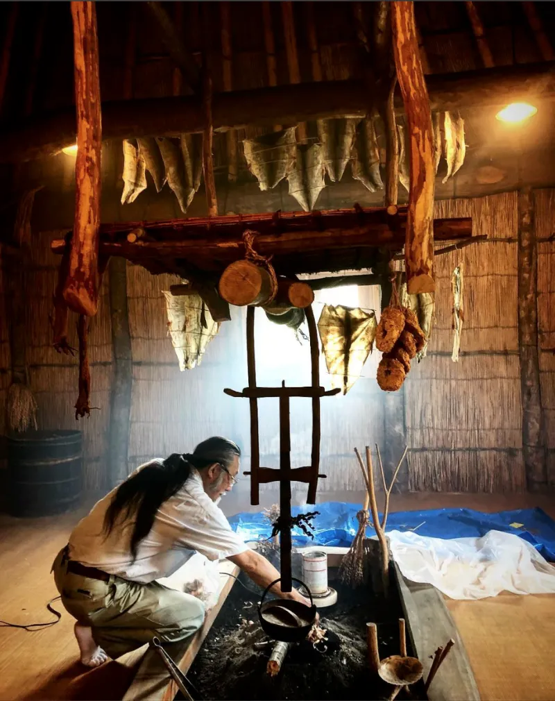 A man, surrounded by fish skins hanging to dry, kneels before a large pit as he demonstrates the tanning process.