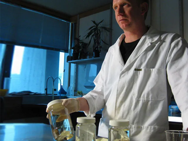 A scientist in a white lab coat stands in laboratory holding a jar of marine specimens. 