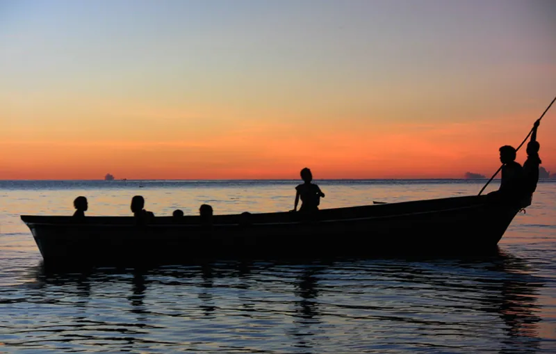 A silhouette of a small boat carrying 8 passengers on tranquil water, with a pink horizon. 