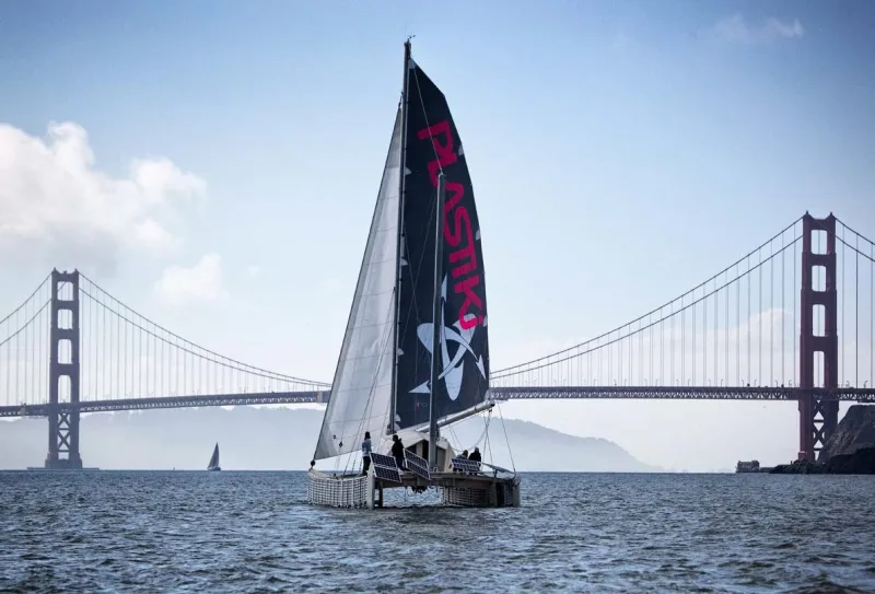 The Plastiki sailing vessel on the water with sails unfurled, in front of a large suspension bridge.