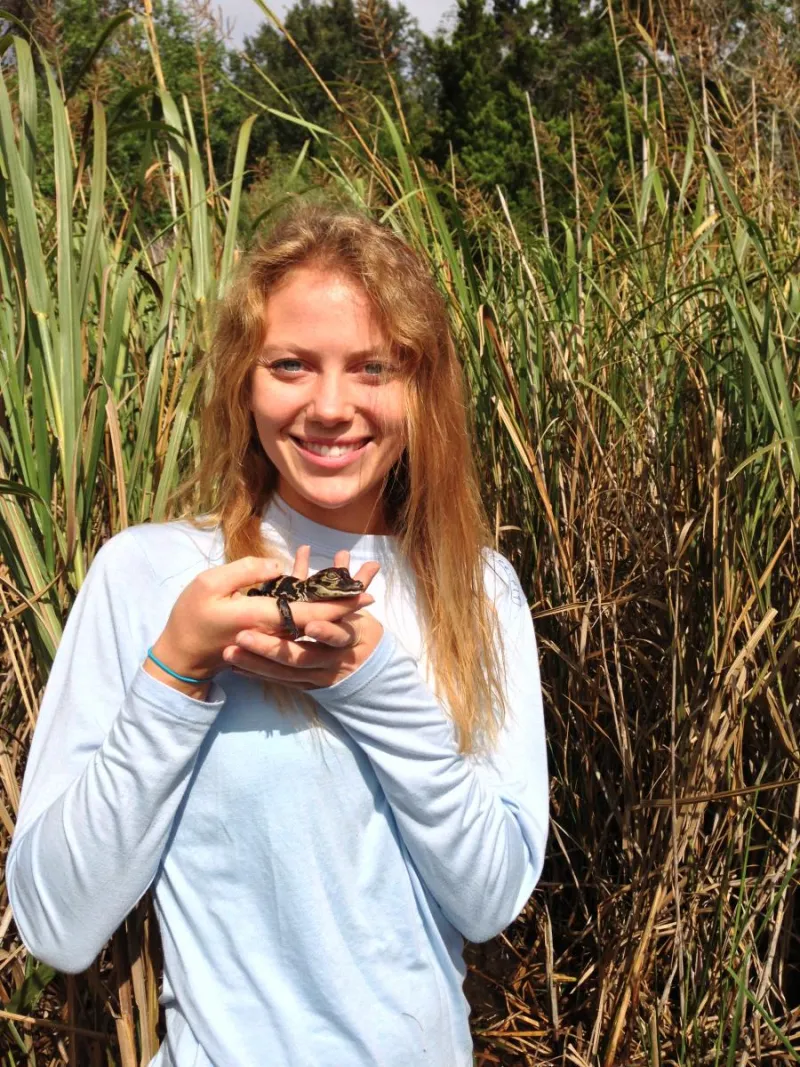 Alexis Temkin stands in marsh grass holding a reptile. 
