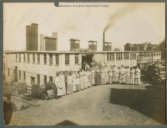 a group of women in period dresses from 1912 stand outside a factory with smoking stacks