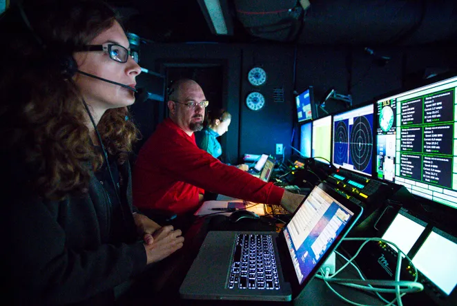 Andrea sits in front of a laptop and huge dashboard of sensors with a headset on.