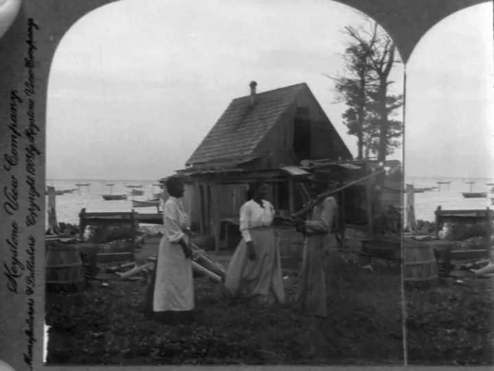A man and two women in long skirts by a small house near the ocean
