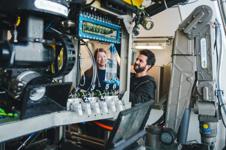 Two researchers hold a plastic bag full of water, surrounded by metal machinery. 