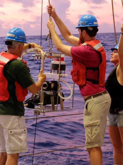 Researchers work with an instrument on a ship while wearing hard hats and life vests.