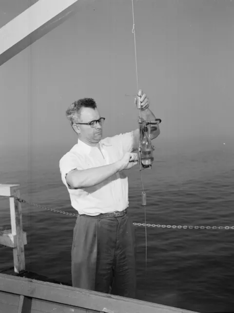 Black and white photo of a man holding up an instrument while on a boat.