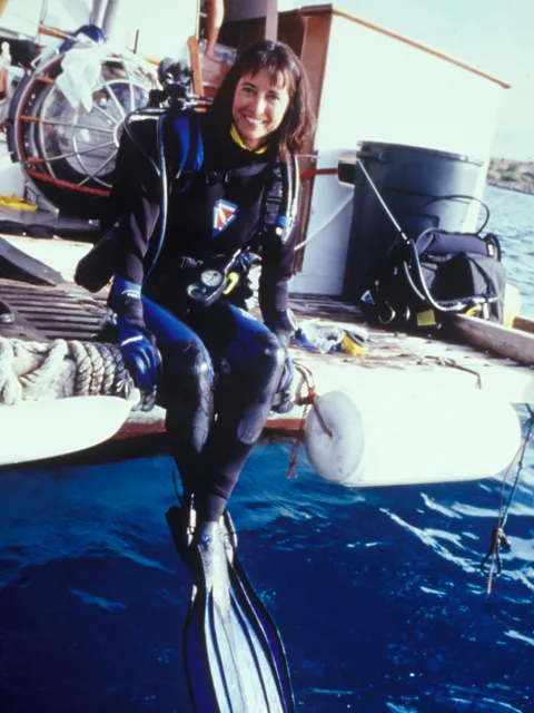 a female scientist with brown hair sits suspended on the back of the boat in a full wetsuit and scuba gear