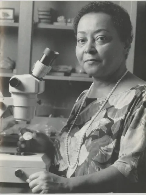a woman with a beaded necklace and floral dress stands by her microscope and a coral specimen in hand