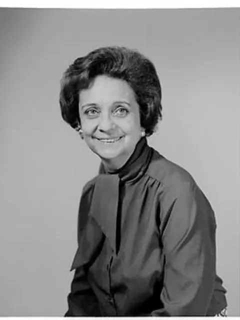 a black and white portrait of a smiling woman with short hair, pearl earrings and a high necked shirt